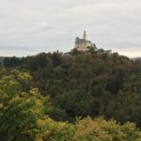 Castel medieval pe o colină împădurită, cu turnuri și ziduri de piatră luminate de soare