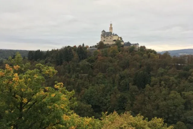 Castel medieval pe o colină împădurită, cu turnuri și ziduri de piatră luminate de soare